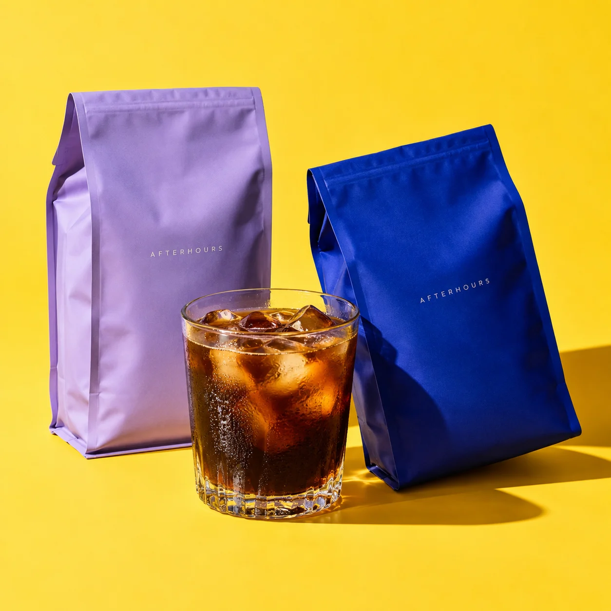 Lavender and cobalt Afterhours specialty coffee bags beside a glass of iced coffee on a yellow studio backdrop